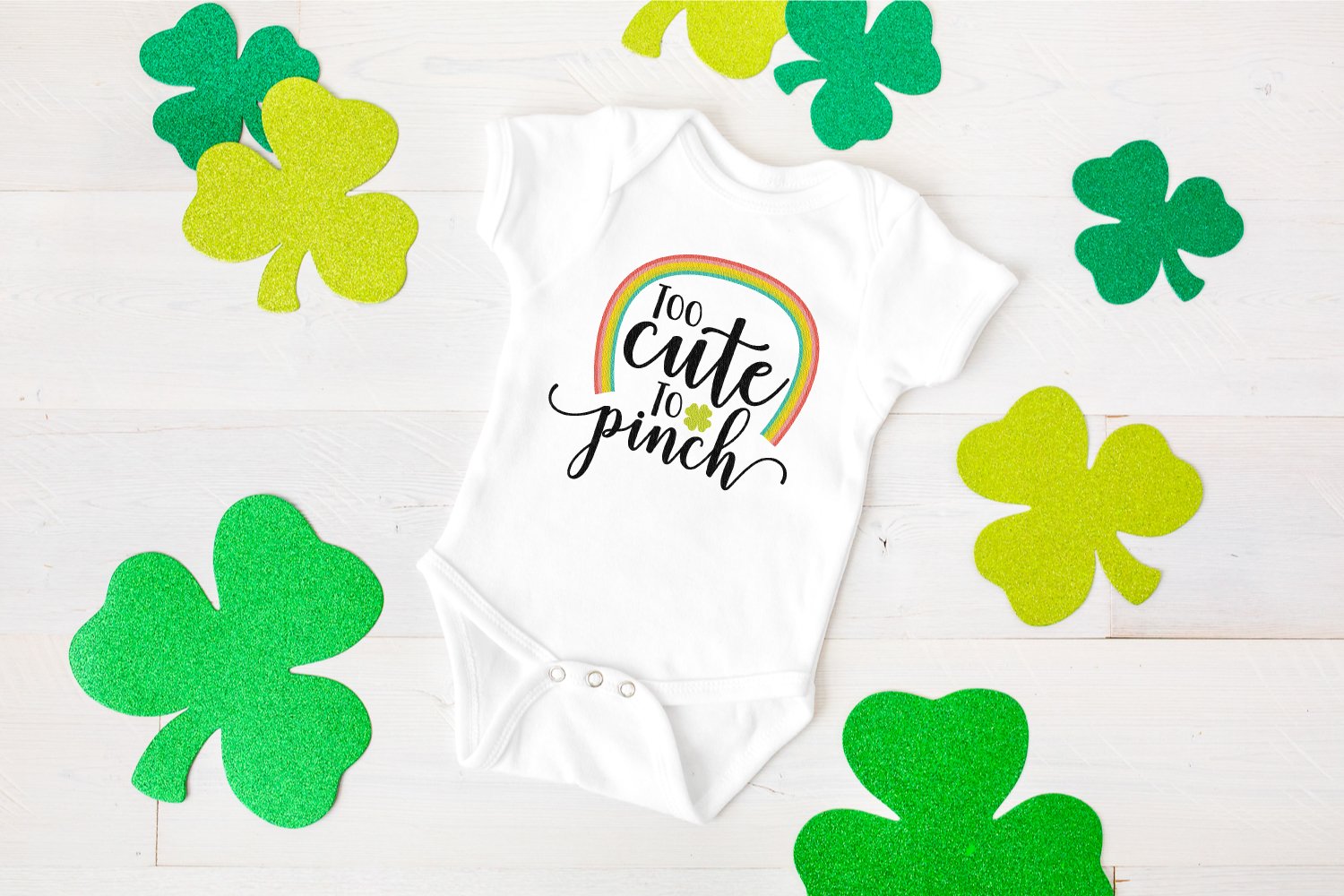 Close up of green shamrocks around a white onesie that is decorated with a rainbow and the saying, \"Too Cute to Pinch\"