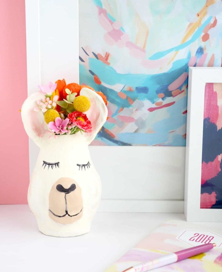 A vase decorated with a llama face with flowers in it sitting on a table next to a couple of paintings