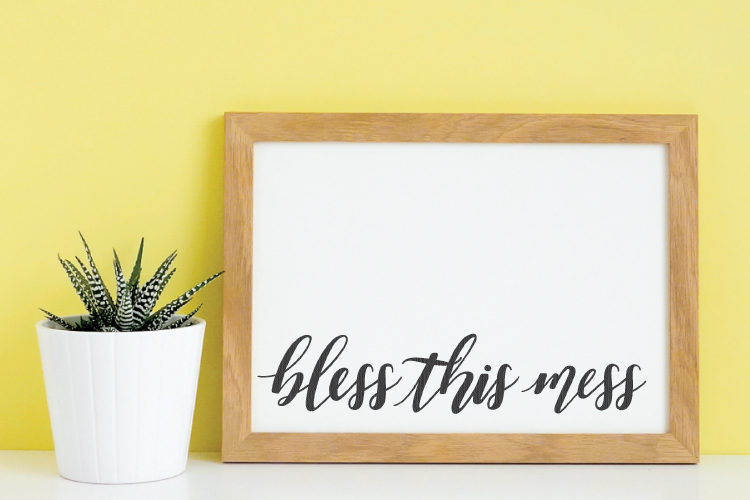 A close up of a potted plant sitting against a yellow wall and next to a wooden framed sign that says, \"Bless this mess\"