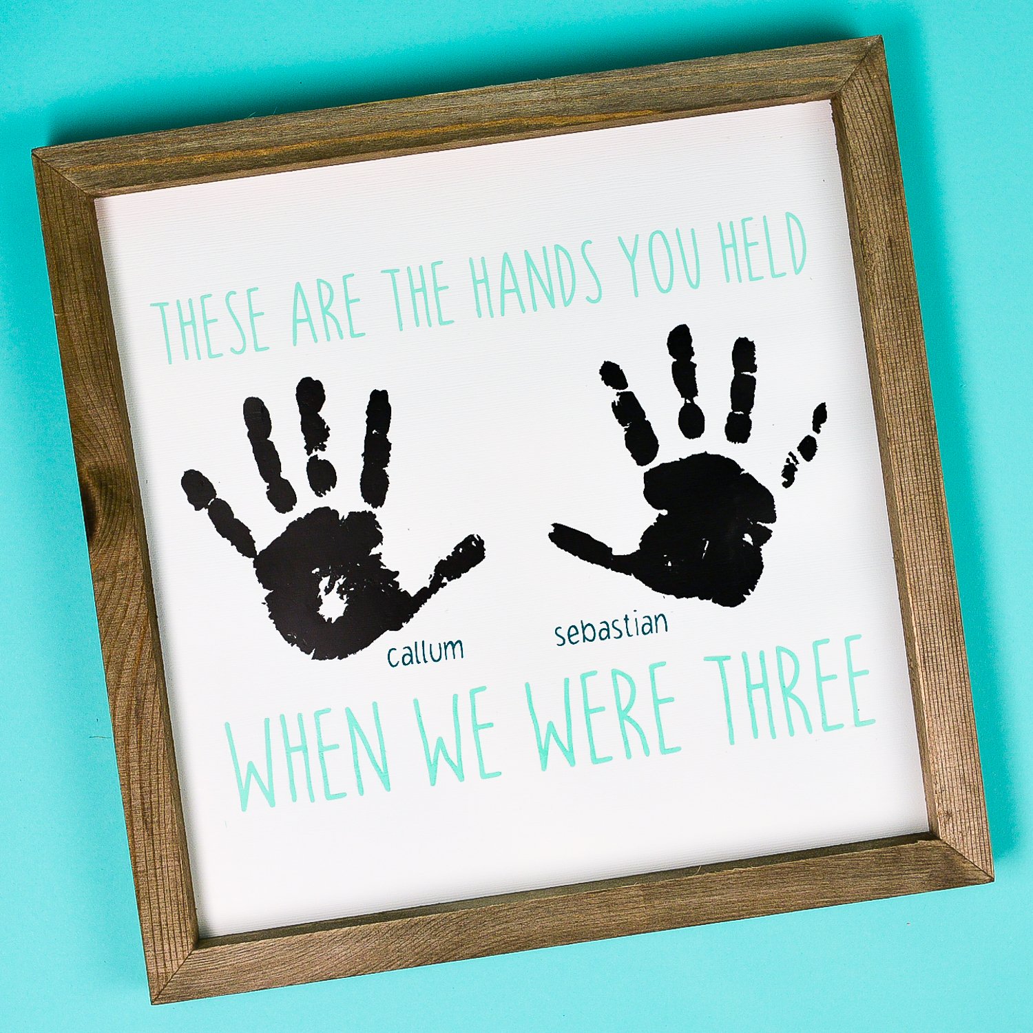 A close up of a wood frame holding a sign that contains two handprints with boy\'s names next to each and says, \"These are the Hand You Held When We Were Three\"