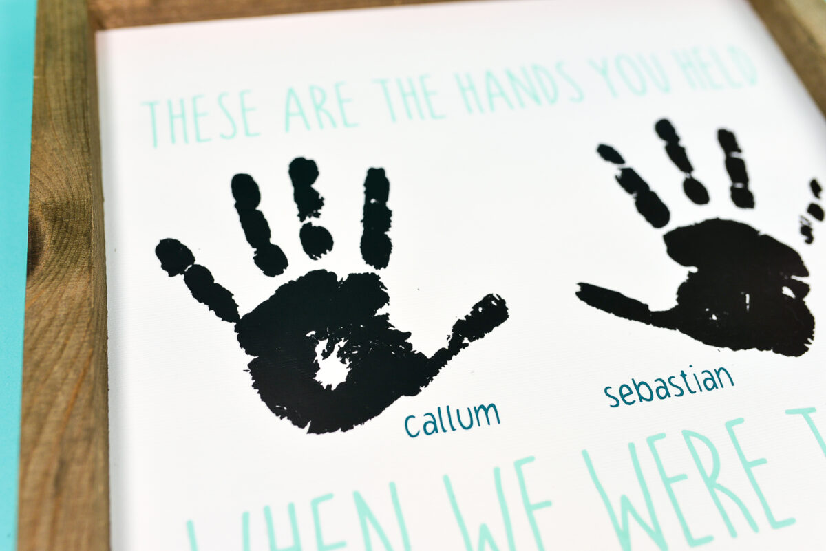 A close up of a wood frame holding a sign that contains two handprints with boy\'s names next to each and says, \"These are the Hand You Held When We Were Three\"