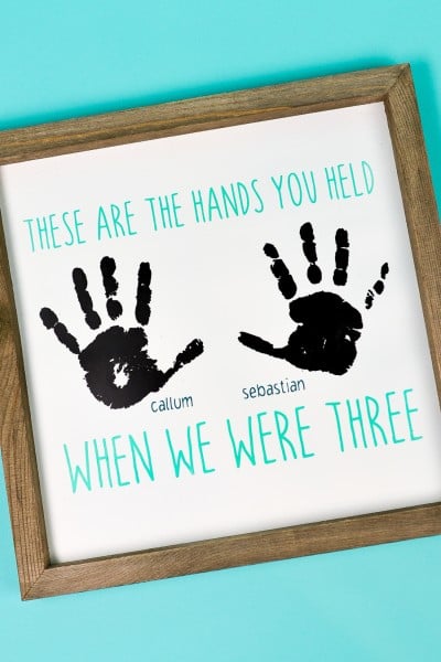 A wood frame holding a sign that contains two handprints with boy's names next to each and says, "These are the Hand You Held When We Were Three"
