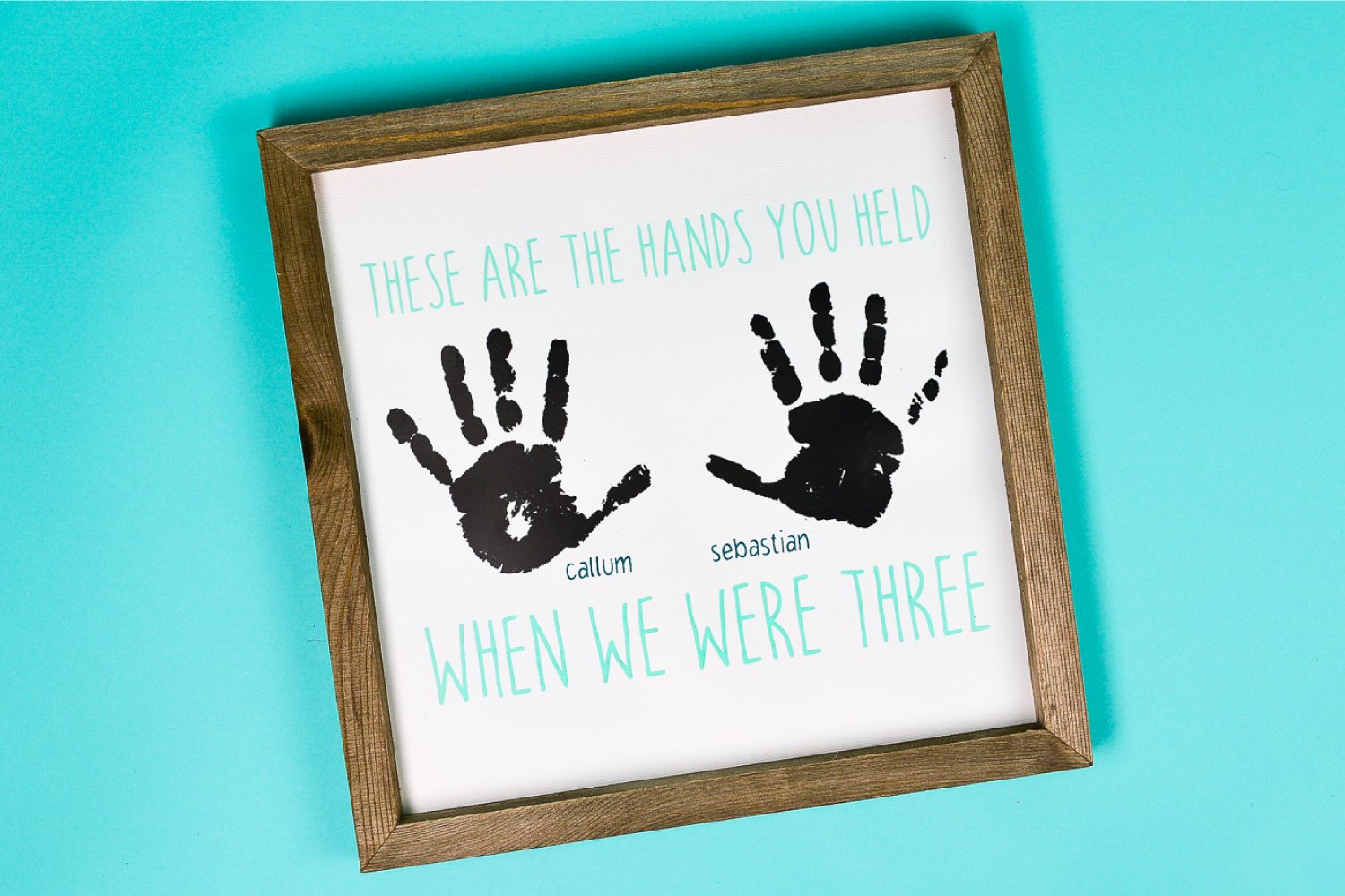 A close up of a wood frame holding a sign that contains two handprints with boy\'s names next to each and says, \"These are the Hand You Held When We Were Three\"