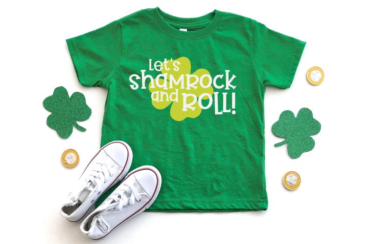 Green shamrocks with a pair of tennis shoes and a green t-shirt decorated with a light green shamrock with the saying, \"Let\'s Shamrock and Roll!\"