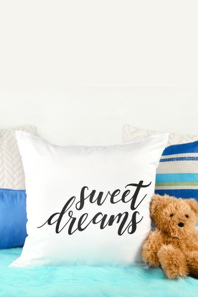 A teddy bear sitting next to several pillows where one pillow is white with the saying, "Sweet Dreams" on it