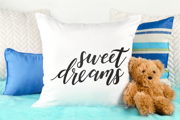 A teddy bear sitting next to several pillows where one pillow is white with the saying, \"Sweet Dreams\" on it