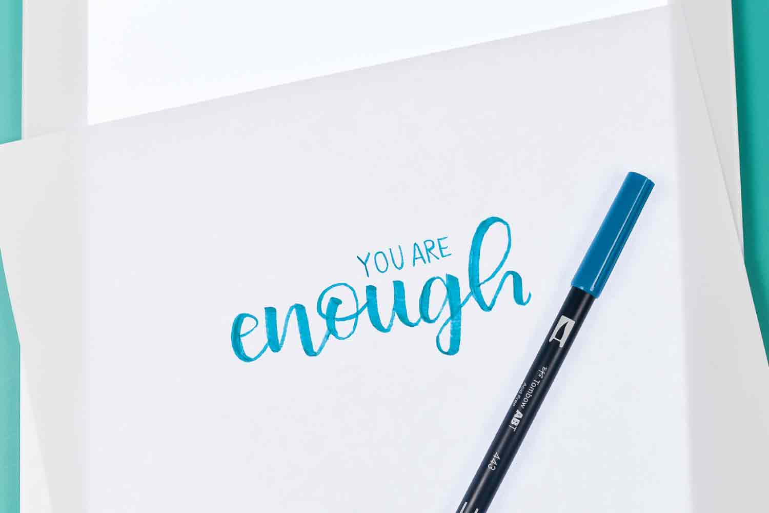 Close up of a Cricut Brightpad with a pen and piece of paper on top of it with the words \'You Are Enough\' written on it