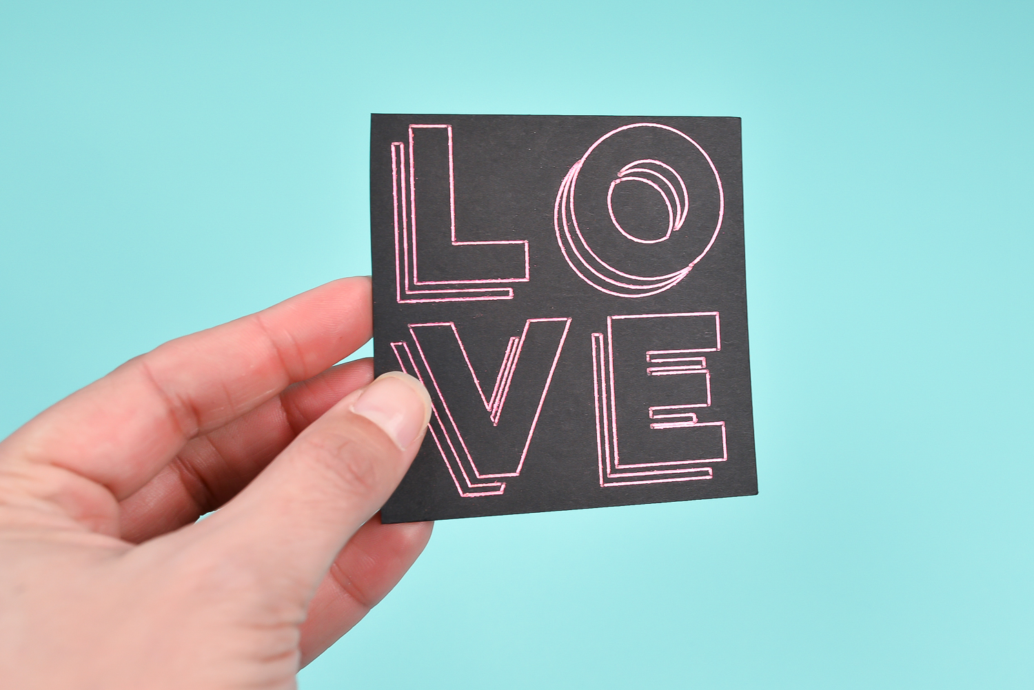 A person holding a piece of black paper that is decorated with foil quill lines that spell out the word \"Love\"