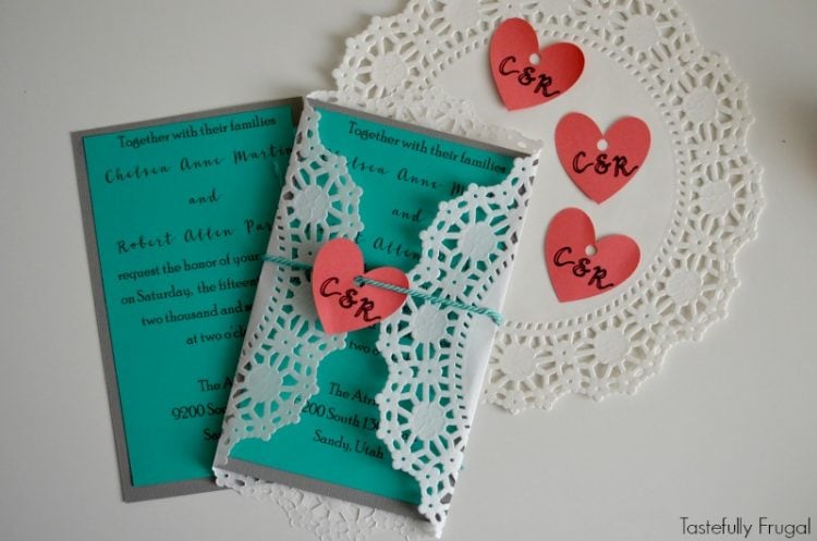Image of two wedding invitations next to a doily and pink monogramed hearts with one wrapped in a paper doily and tied with string and the pink paper monogrammed heart attached