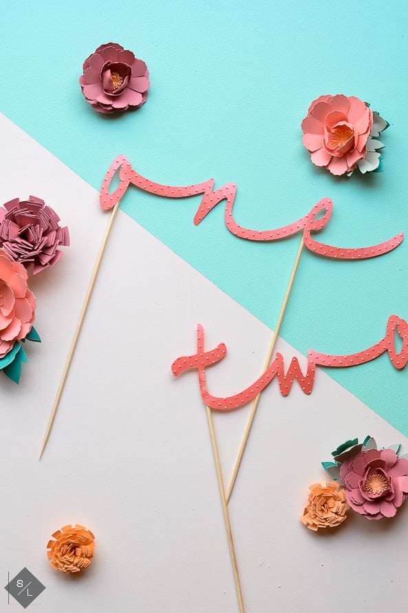 Paper cut flowers on a table along with the words \'one\' and \'two\' cut out of paper and with toothpicks attached to the words