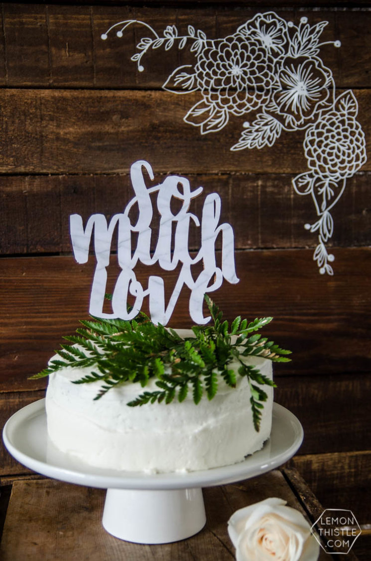 A wooden table that holds a single white rose and a white cake decorated with greenery and a wedding cake topper that says, \"So Much Love\"