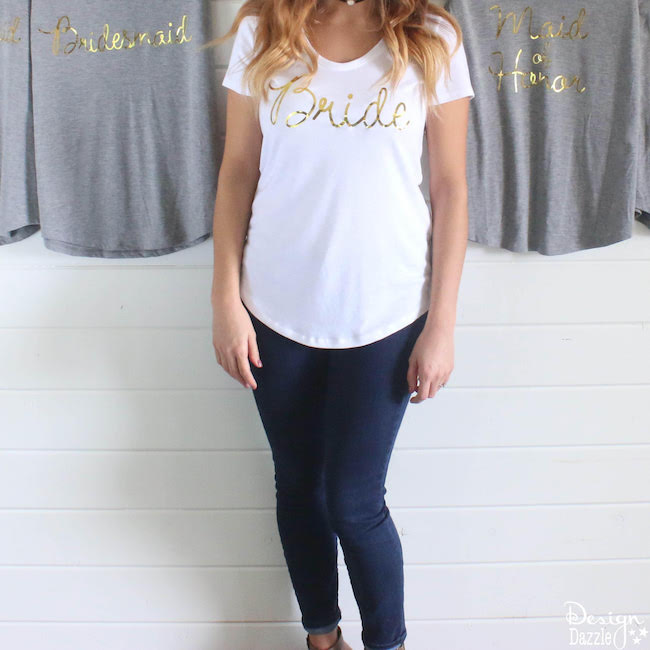 A woman dressed in black leggings and a white t-shirt that says, \"Bride\" and two other gray shirts hanging behind her that say, \"Bridesmaid\" and \"Maid of Honor\"