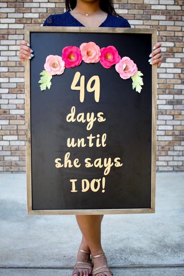 A woman standing next to a brick wall and holding a chalkboard decorated with paper cut flowers and the saying, \"49 days until she says I DO!\"