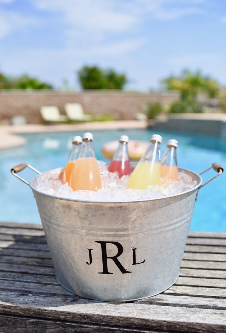 A monogrammed tin ice bucket filled with beverages and sitting on a deck next to a swimming pool