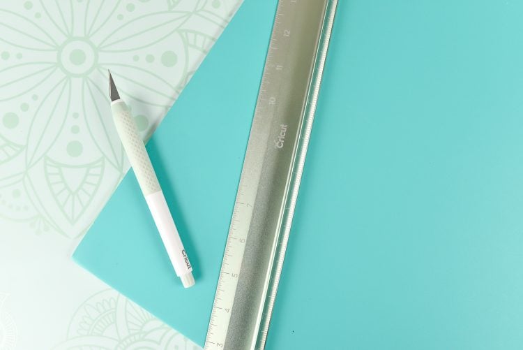 A ruler and the Cricut True Control Knife lying on top of an aqua colored piece of fabric and the Cricut self-healing mat