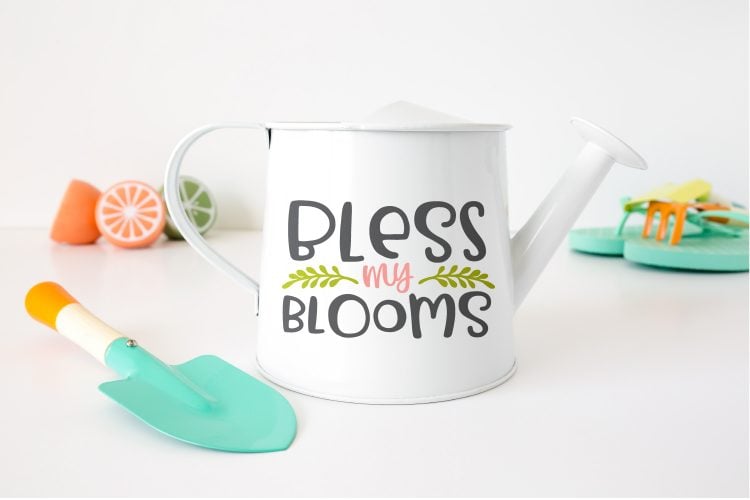 Close up of a white watering can with the saying, \"Bless My Blooms\" and a pair of sandals, a small gardening scoop and sliced fruit