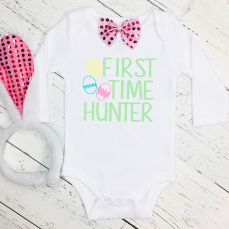 White onesie decorated with a pink bow tie and Easter eggs with wording First Time Hunter