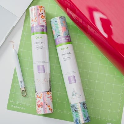Partial picture of a red Cricut machine and a laptop along with a Cricut green map, a Cricut weeding tool and two rolls of patterned iron on vinyl