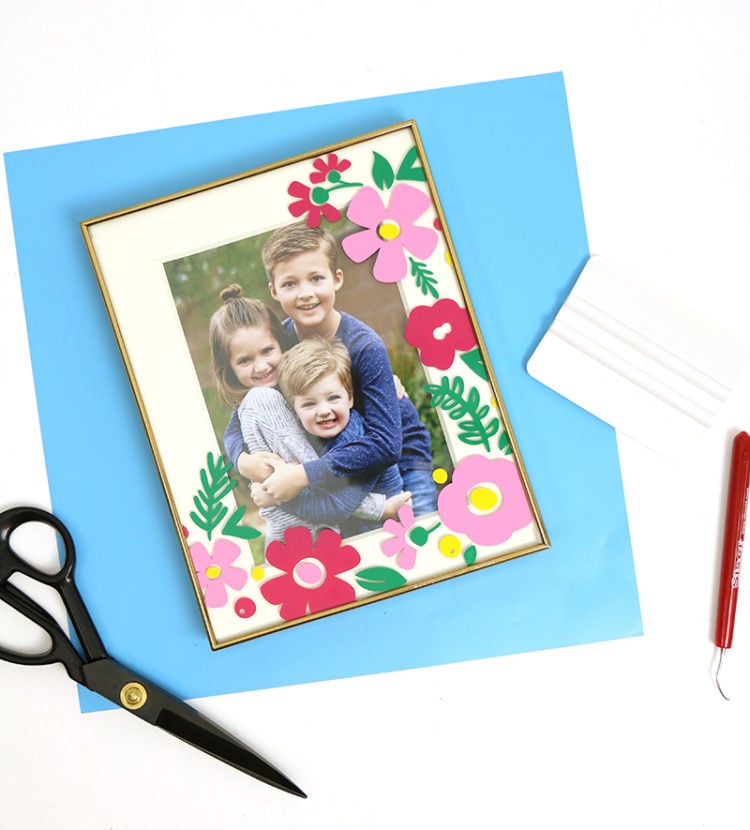 A scissors, a weeding tool and a framed picture of three young children with the mat around the picture decorated with paper cut flowers and greenery
