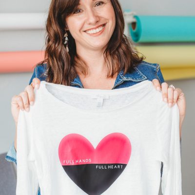Cori George holding up a long-sleeved white shirt with a heart image on it that says, "Full Hands, Full Heart"