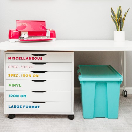 An aqua blue tote under a table that has a Cricut machine on top of it and a plant.  The table has drawer's underneath that are labeled as "Miscellaneous", "Spec Vinyl", "Spec Iron On", "Vinyl" and "Iron On" and "Large Format"