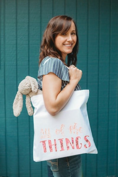 Cori George holding a tote bag with a stuffed toy bunny hanging out of it and the tote has the saying, "ALL of the Things"