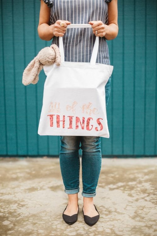 A woman holding a holding a tote bag with a stuffed toy bunny hanging out of it and the tote has the saying, "ALL of the Things"