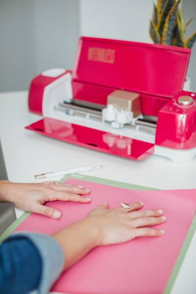 Red Cricut machine and a woman's hands applying a piece of pink vinyl to a green Cricut mat