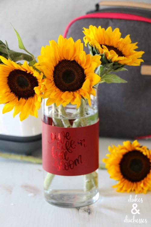 Sunflowers in a vase that is wrapped with a leather vase wrap that says, \"Live Life in Full Bloom\"