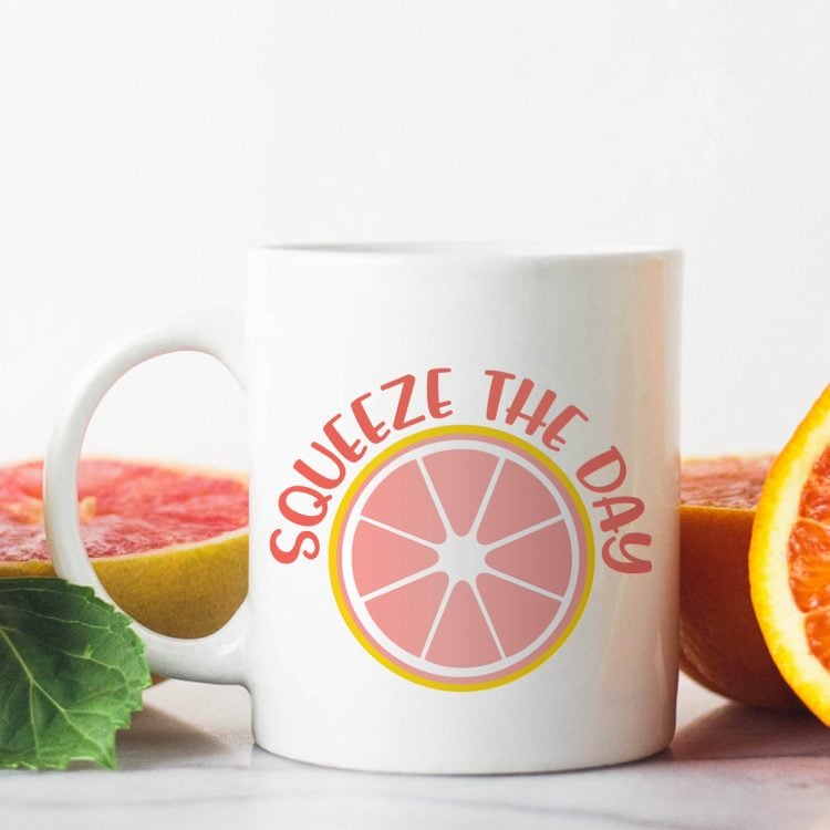 A grapefruit cut in half and a cup of coffee that has an image of a half of a grapefruit and the saying, \'Squeeze the Day\"