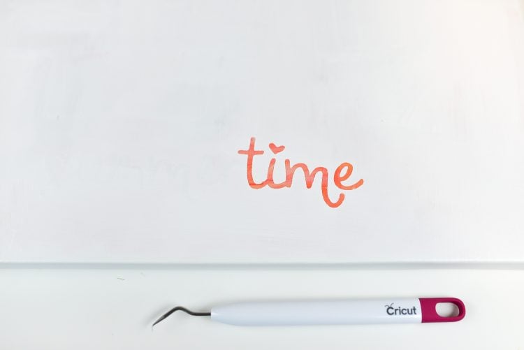 A Crciut weeding tool lying next to a white canvas with the word 'time' displaying