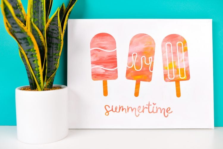 A plant in a white pot sitting next to a white canvas decorated with three popsicle images and the word summertime