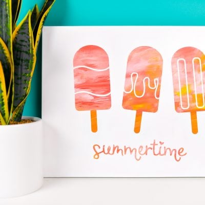 A plant in a white pot sitting next to a white canvas decorated with three popsicle images and the word summertime