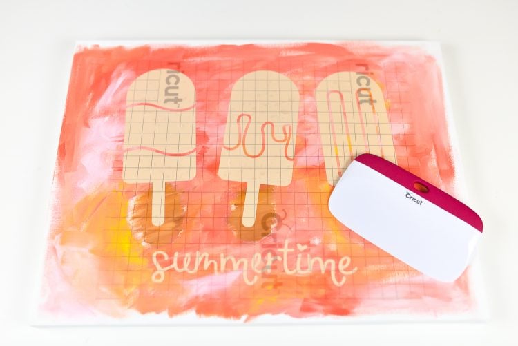A cricut brayer sitting on top of a cut file pattern of three popsicles and the word summertime laying over top of a painted canvas