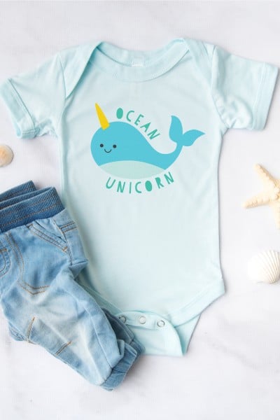 Seashells surrounding a pair of baby blue jeans and a light blue onesie that is decorated with a blue whale with a unicorn horn with the saying, "Ocean Unicorn"
