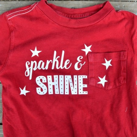 A red t-shirt lying on a wooden table and is decorated with white stars and says, "Sparkle & Shine"