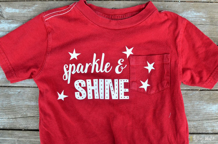 A red t-shirt lying on a wooden table and is decorated with white stars and says, "Sparkle & Shine"