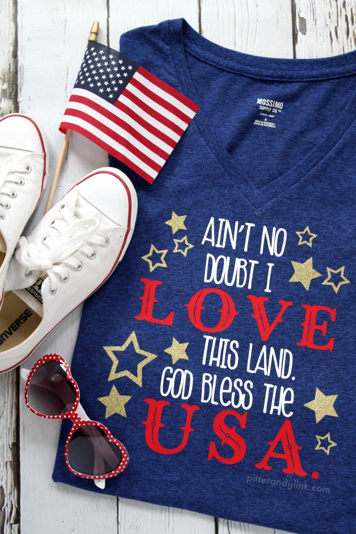 A pair of heart-shaped red sunglasses, a pair of tennis shoes, a USA flag pennant and a dark blue t-shirt that says, "Ain't No Doubt I Love This Land.  God Bless the USA"