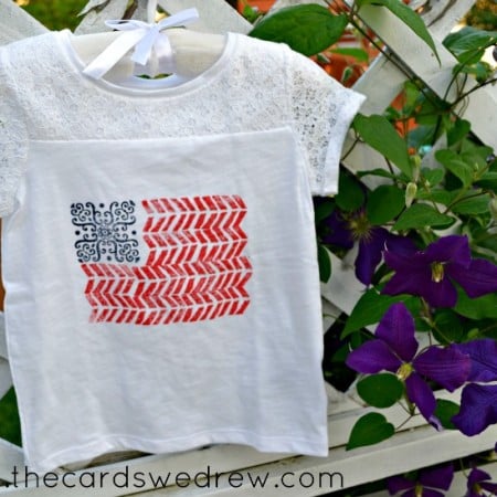 A white lace topped t-shirt with an image on it that resembles the flag of the United States of America hanging on a hanger on a trellis next to some flowering vine