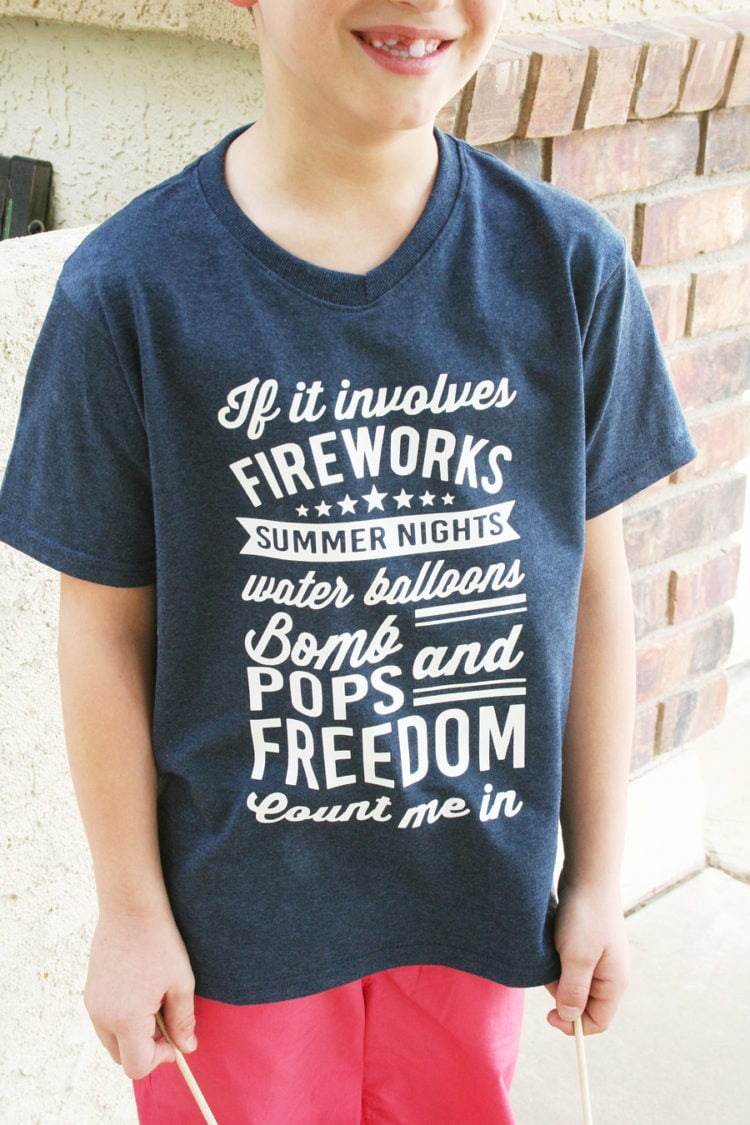 A young boy standing by a brick wall, wearing red shorts and a blue t-shirt that says, "If it involves fireworks, summer nights, water balloons, bomb pops and freedom, count me in"
