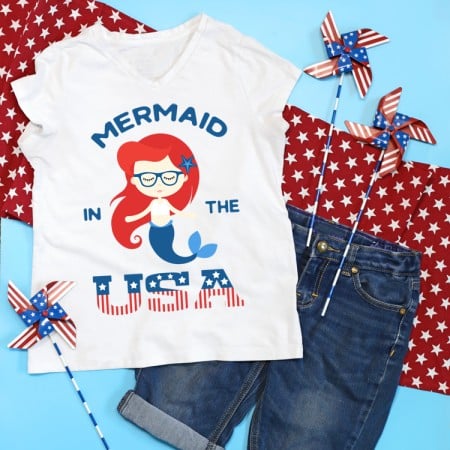 A red piece of material with white stars on it surrounded by three American flag pinwheels, a pair of blue jean shorts and a white t-shirt decorated with a mermaid that says, "Mermaid in the USA"