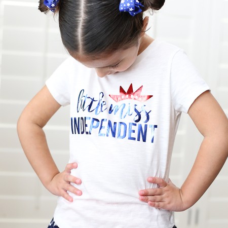 A girl wearing a blue skirt with white stars on it and a white t-shirt that says, "Little Miss Independent"