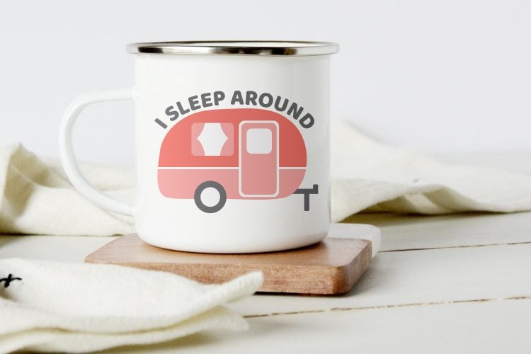 A coffee mug sitting on a coaster and the mug is decorated with an old-style camper with the saying, \"I Sleep Around\"