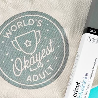 A box of Cricut Infusible Ink lying on top of a canvas style tote that says, "World's Okayest Adult"
