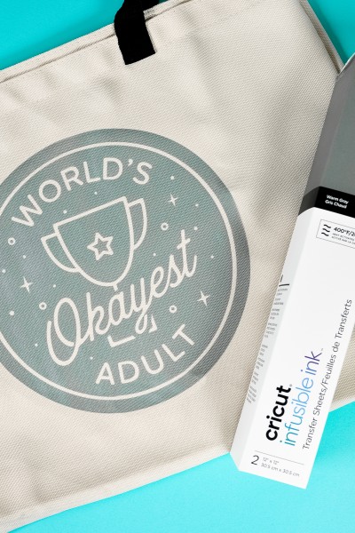 A box of Cricut Infusible Ink lying on top of a canvas style tote that says, "World's Okayest Adult"