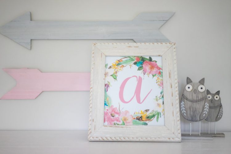 A gray and pink wooden arrow hanging on a wall and underneath is a picture of the letter \"a\" in a white frame alongside of two wooden figures of an owl