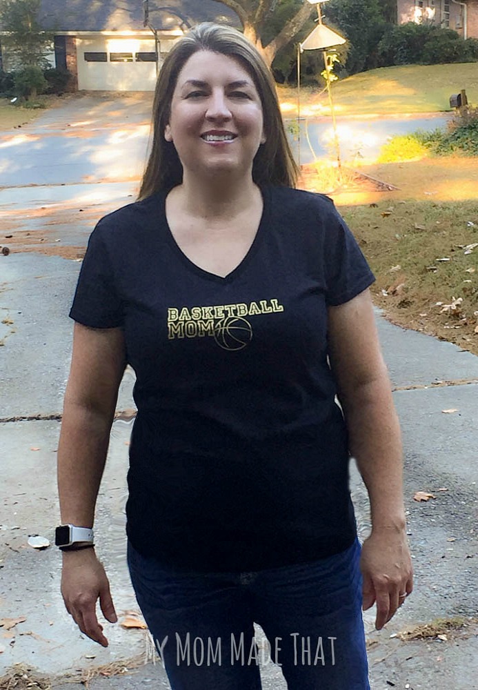 A lady wearing a black V-neck t-shirt with the saying, \"Basketball Mom\" and an image of a basketball in gold vinyl