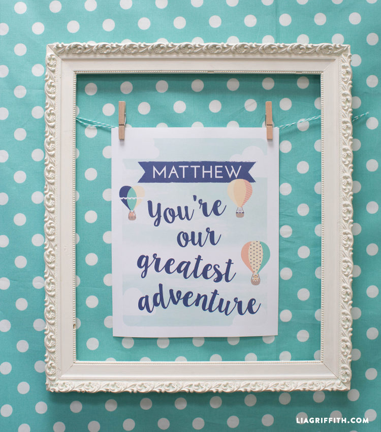 An aqua colored wall with white polka dots all over it and a white frame hanging on the wall with a sheet of paper hung by tiny clothespins and the paper is decorated with hot air balloons and a personalized name with the saying, \"You\'re Our Greatest Adventure\"