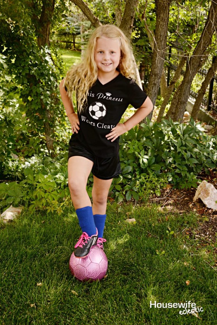 A young girl standing next to a tree wearing a black uniform and she has her foot on a pink soccer ball.  The black shirt has an image of a soccer ball on it and the saying \"This Princess Wears Cleats\"