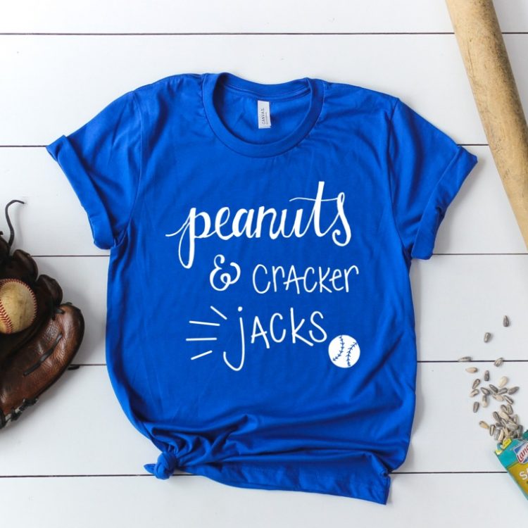 A blue shirt decorated with the saying, \"Peanuts & Cracker Jacks\" next to a baseball in a mitt, a baseball bat a package of sunflower seeds